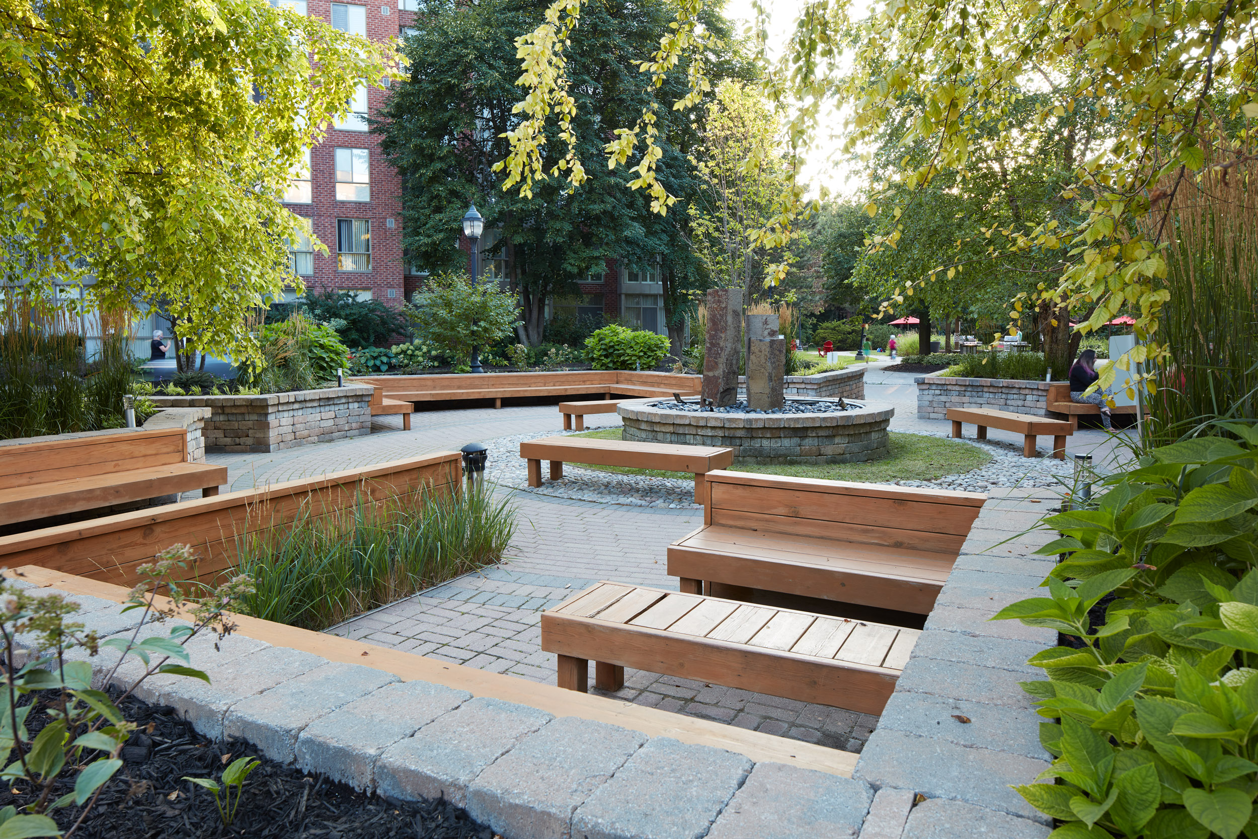 This image shows a serene courtyard or garden area, thoughtfully designed with multiple wooden benches arranged around a circular stone fountain. The setting is peaceful, featuring lush greenery, including trees and manicured plants, and brick paving. The overall style is modern and minimalist with a calming atmosphere. In the background, a residential building is partially visible. A few people are subtly visible in the background, suggesting a community or communal space.