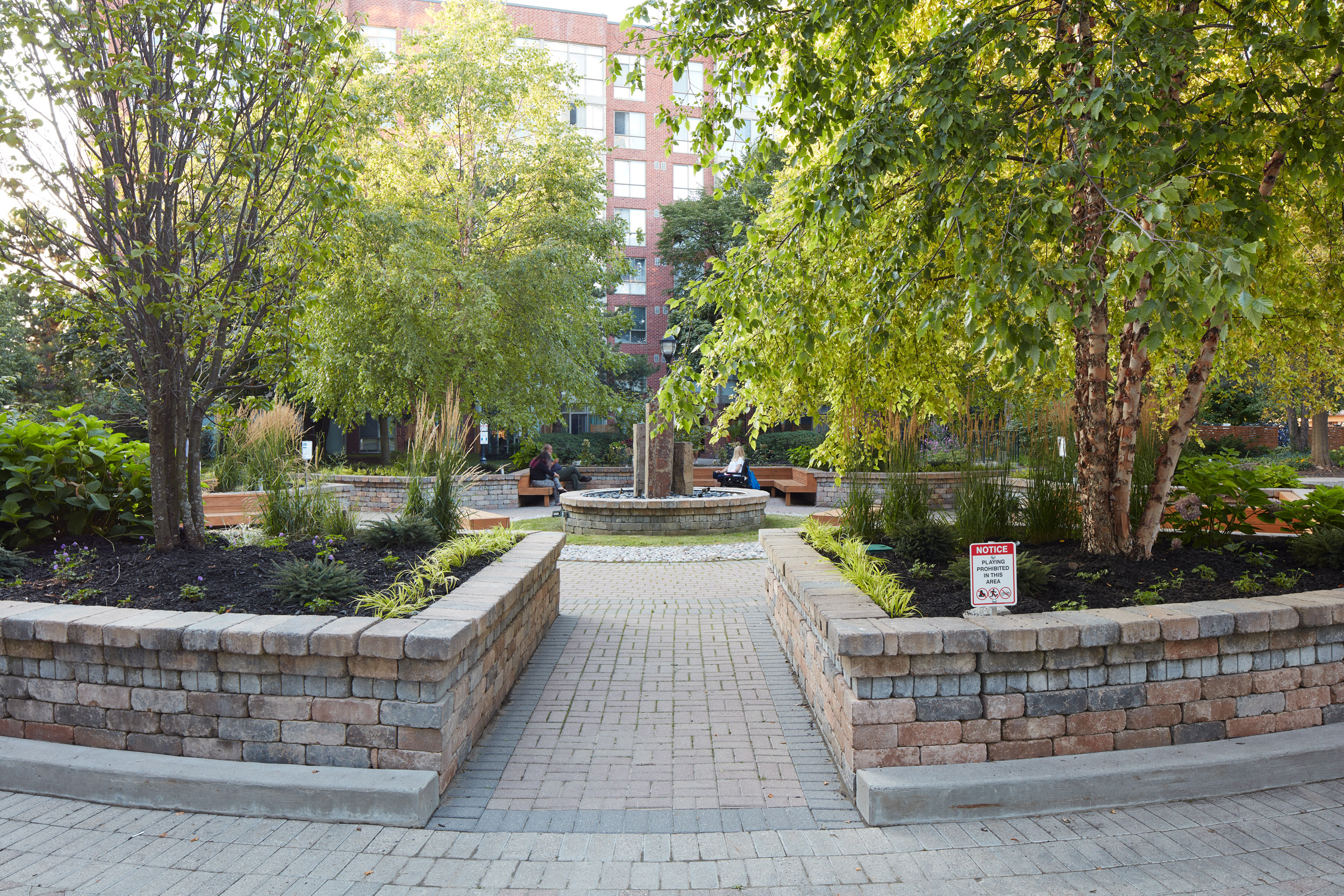 Stone Retaining walls flank entranceway to garden communal area. Garden beds contain trees and perennials. 