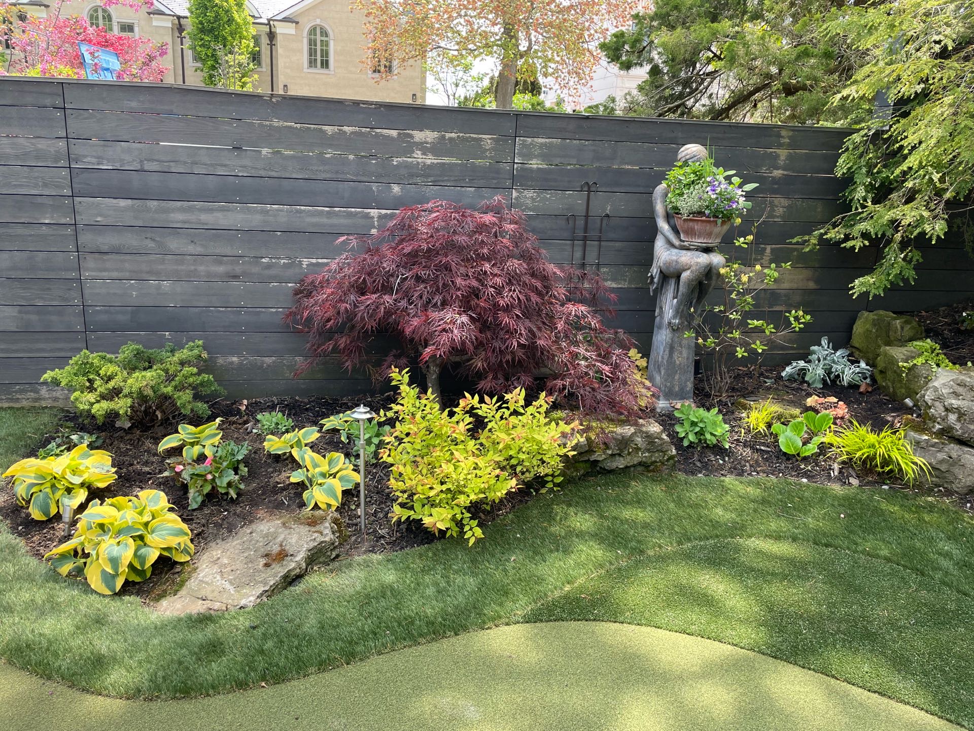 Backyard fence with perennials planted along the base. Outdoor statue stands in the back